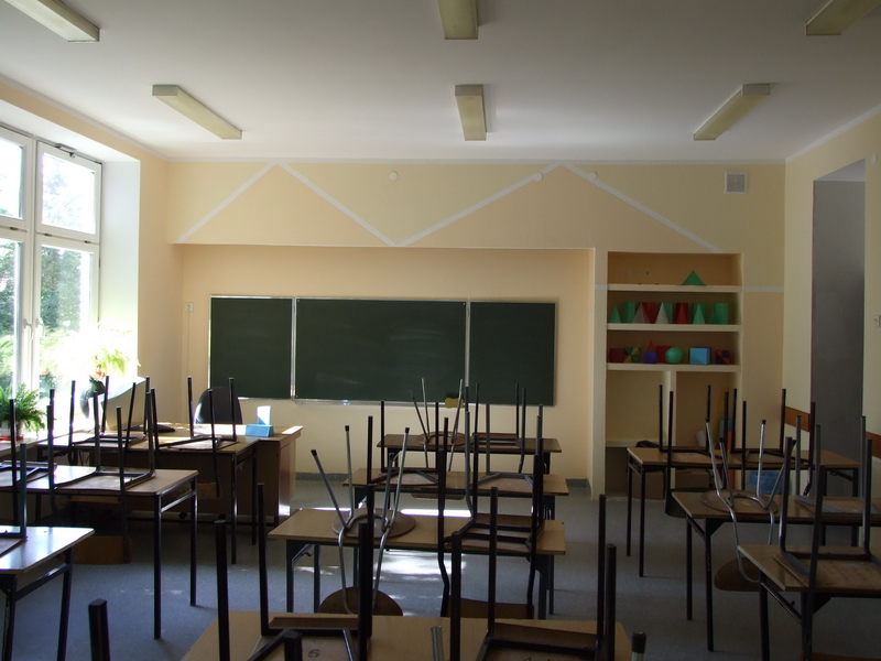 Pusta klasa szkolna z krzesłami odwróconymi na biurkach, zielona tablica i geometryczne bryły na półce, świeżo pomalowane ściany w odcieniach beżu.